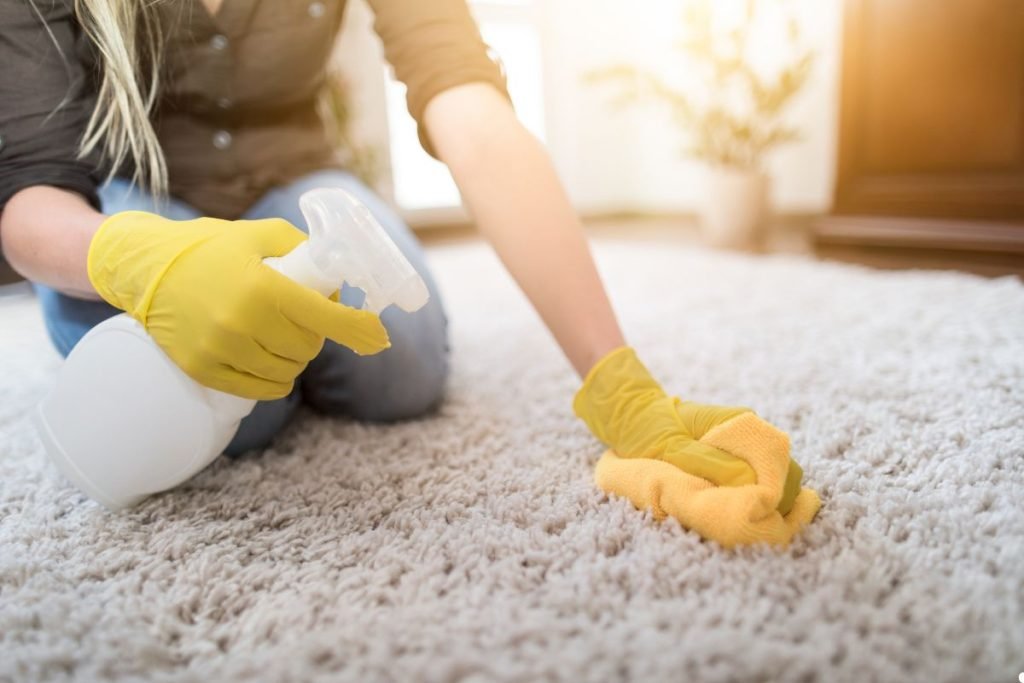 A woman wearing a yellow glove cleans a carpet, focusing on removing dirt and stains with a cleaning tool.